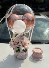 Pink-themed Mother’s Day gift setup with decorative balloons, a flower bouquet, a customized cake in a lighted transparent box, chocolates