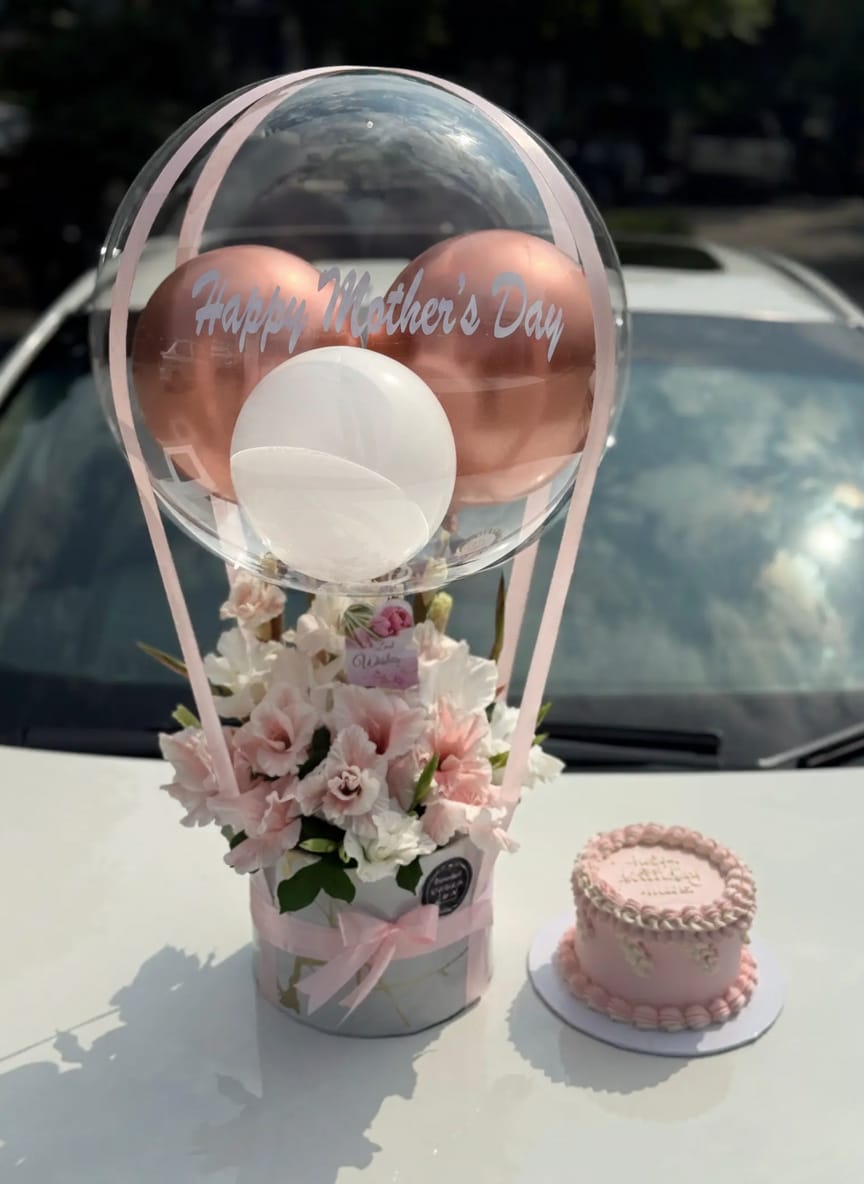 Pink-themed Mother’s Day gift setup with decorative balloons, a flower bouquet, a customized cake in a lighted transparent box, chocolates