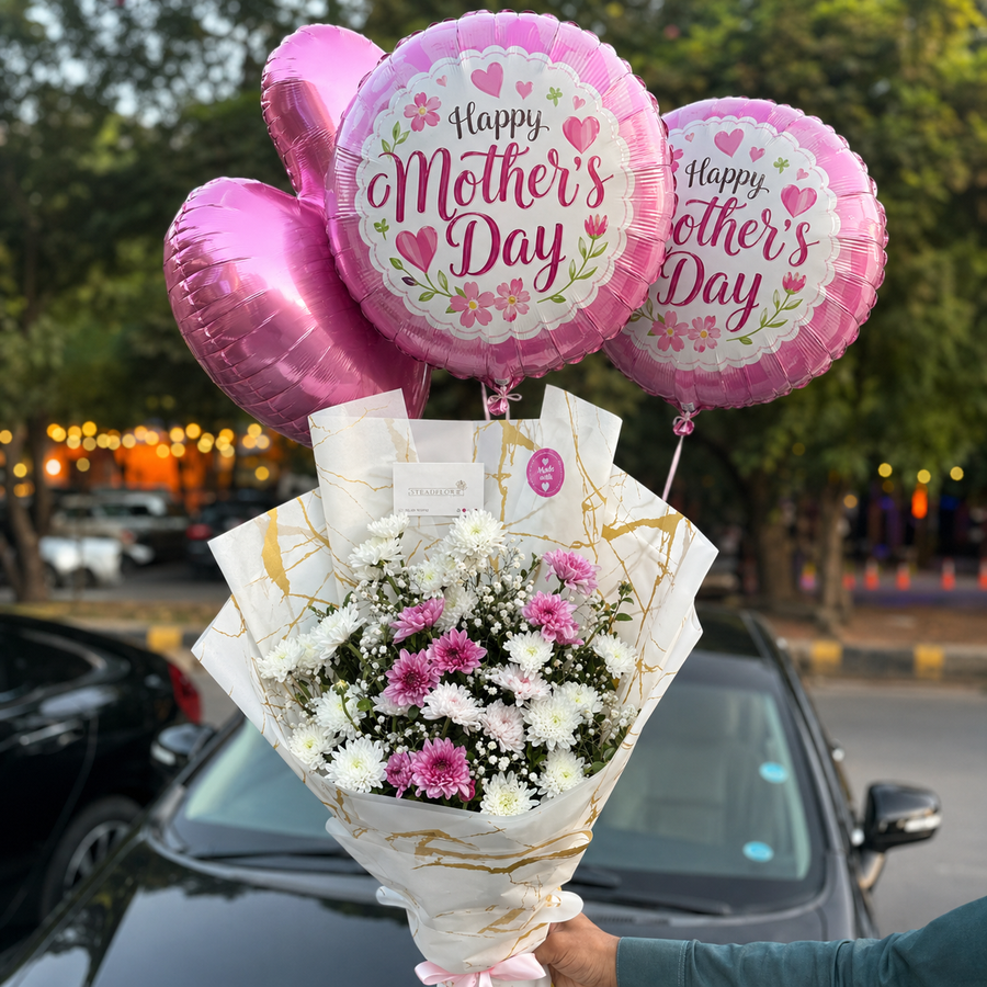 Mother’s Day flower bouquet with pink Happy Mother’s Day balloons, pink heart balloon, white and pink flowers, gold marble wrapping paper, and pink ribbon, held outdoors in front of a car.