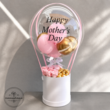 Decorative balloon arrangement with 'Happy Mother's Day' text, pink and gold balloons, and flowers in a white box on a gray background.