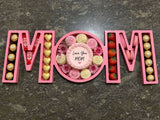 A wooden tray engraved with “MOM,” filled with strawberry macarons and Ferrero Rocher chocolates, arranged as a sweet and elegant gift for mom.