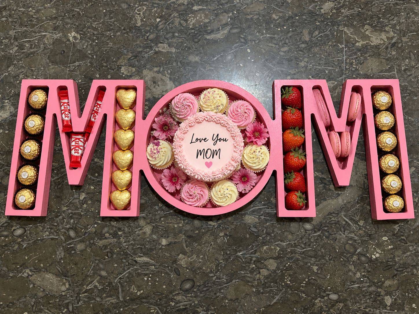 A wooden tray engraved with “MOM,” filled with strawberry macarons and Ferrero Rocher chocolates, arranged as a sweet and elegant gift for mom.