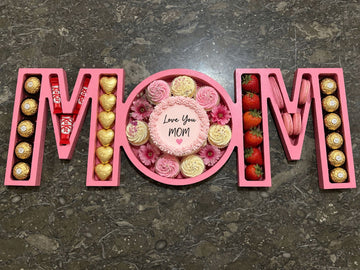A wooden tray engraved with “MOM,” filled with strawberry macarons and Ferrero Rocher chocolates, arranged as a sweet and elegant gift for mom.