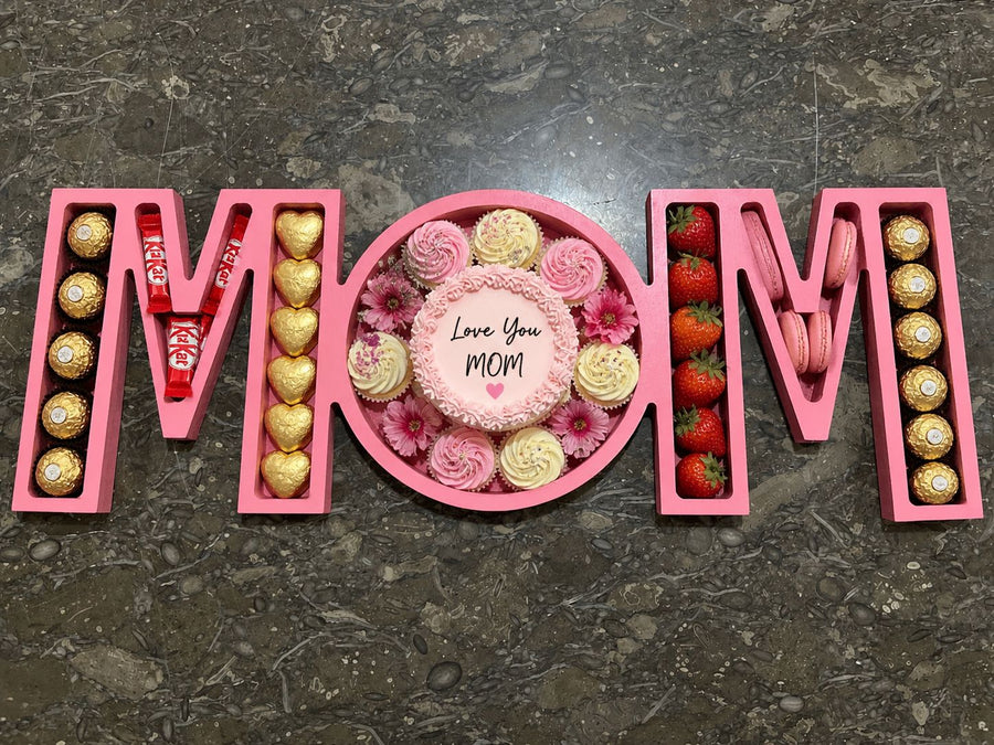 A wooden tray engraved with “MOM,” filled with strawberry macarons and Ferrero Rocher chocolates, arranged as a sweet and elegant gift for mom.