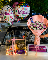 A Mother’s Day gift arrangement featuring colorful “Happy Mother’s Day” balloons, a bouquet of pink and white flowers, a chocolate cake in a lit display box, chocolates, and a wrapped gift box