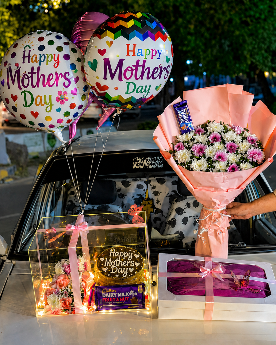 A Mother’s Day gift arrangement featuring colorful “Happy Mother’s Day” balloons, a bouquet of pink and white flowers, a chocolate cake in a lit display box, chocolates, and a wrapped gift box