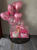 A Mother’s Day gift arrangement placed on a small table, featuring a transparent box with a cake and pink roses inside, decorated with a pink ribbon, warm lights, and a bouquet of heart-shaped and confetti balloons above.