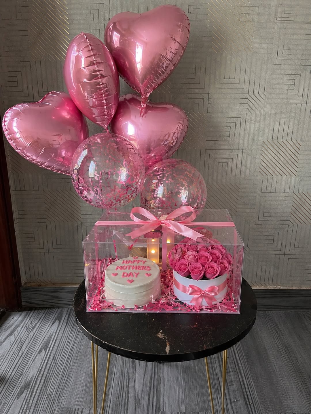 A Mother’s Day gift arrangement placed on a small table, featuring a transparent box with a cake and pink roses inside, decorated with a pink ribbon, warm lights, and a bouquet of heart-shaped and confetti balloons above.