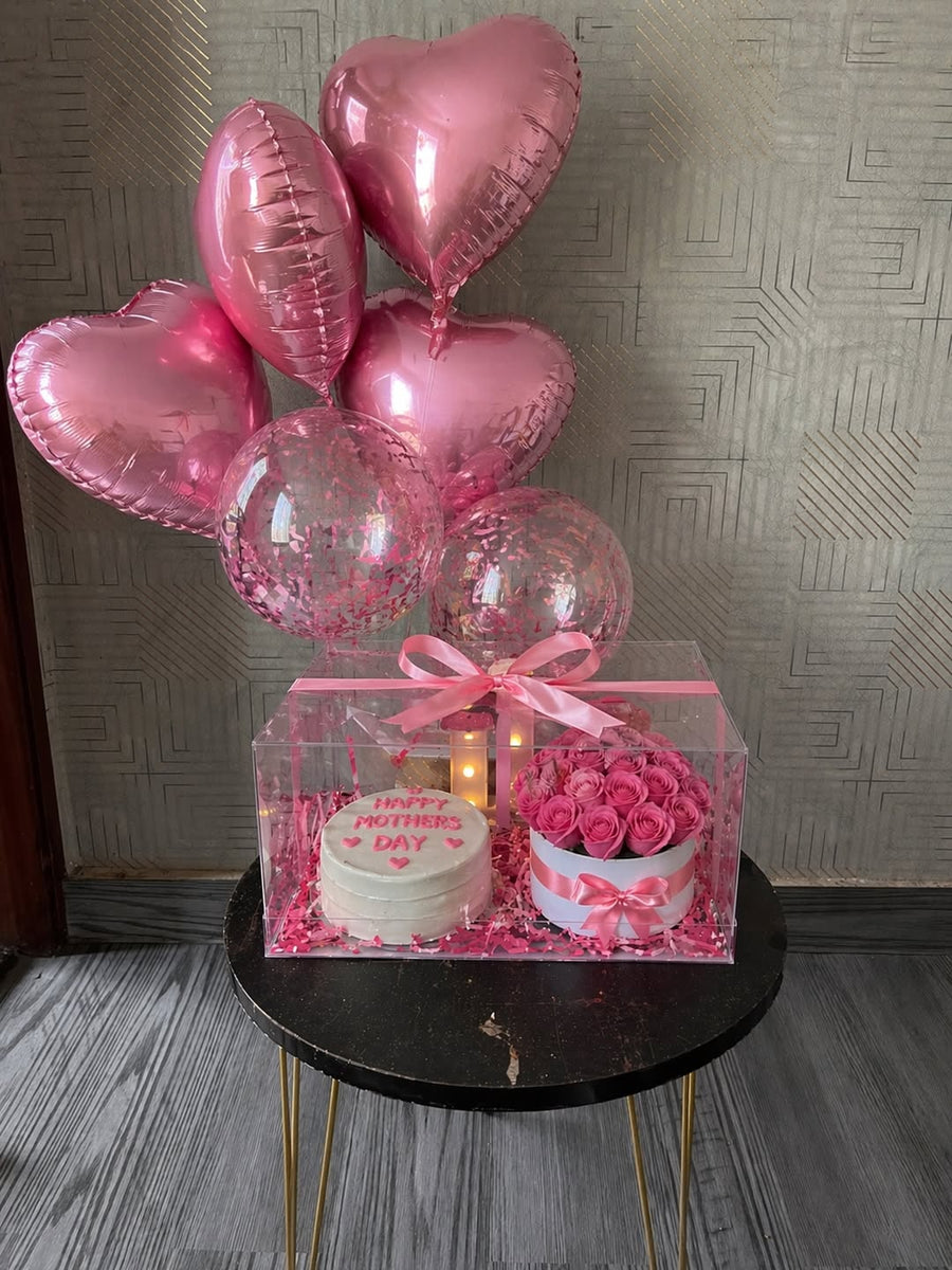 A Mother’s Day gift arrangement placed on a small table, featuring a transparent box with a cake and pink roses inside, decorated with a pink ribbon, warm lights, and a bouquet of heart-shaped and confetti balloons above.