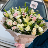 Mother’s Day pastel flower bouquet with white lilies, pink roses, chrysanthemums, baby’s breath, pink marble wrapping, ribbon bow, and Happy Mother’s Day topper held in front of a car.