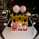 Mother’s Day luxury gift box with gold MOM balloons, Happy Mother’s Day balloons, teddy bear, pink flowers, cake, chocolates, fairy lights, and ribbon displayed on a white car hood at night.